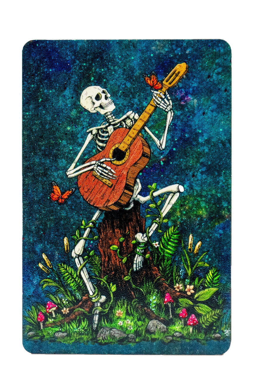 Magnet showing a skeleton playing acoustic guitar with butterflies, surrounded by flowers and vines, sitting on a tree stump. Blue mottled background.