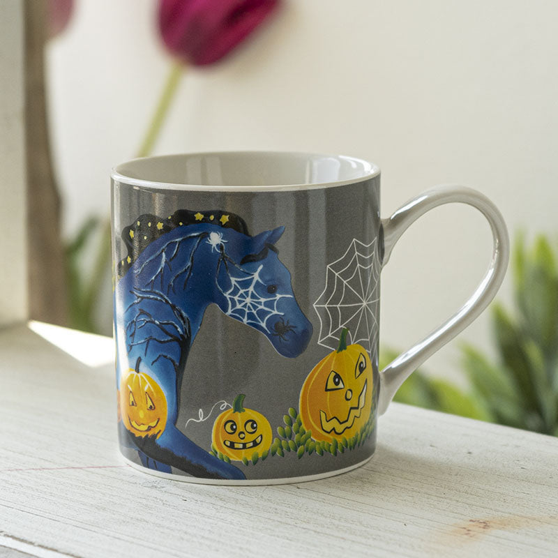 A gray ceramic mug with a white handle and interior, decorated with a Halloween design featuring a blue horse with spiderwebs, orange jack-o'-lantern pumpkins, white cobwebs, and other spooky details