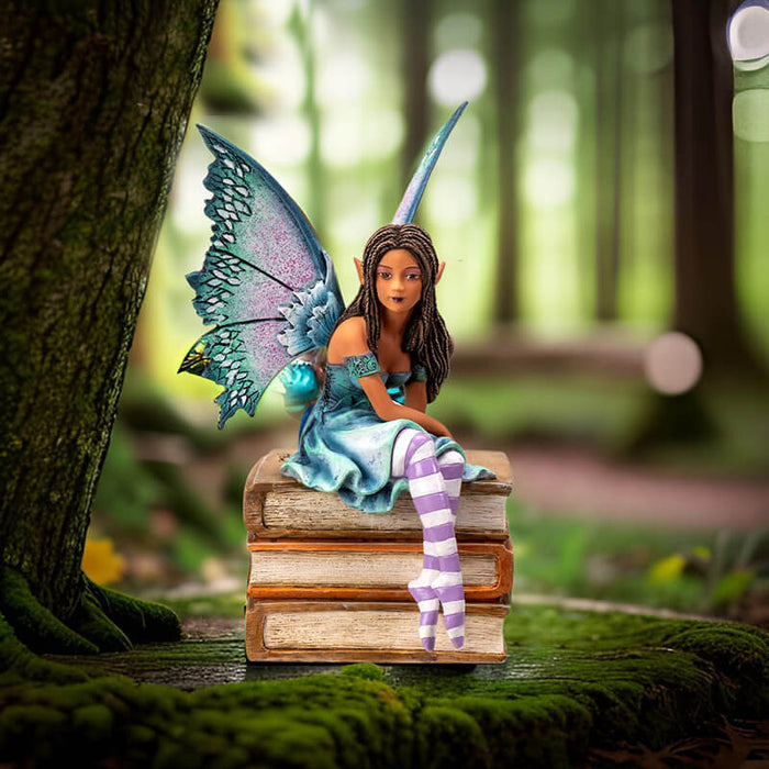 Figurine of a tanned skin fairy with brown braided hair and teal-purple wings in a blue-green dress, perched on a stack of tan books. Striped white and purple stockings.
