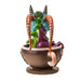 Figurine of a blue, green and purple dragon with red horns popping out from a faux mug of hot chocolate with white writing on brown. Back view