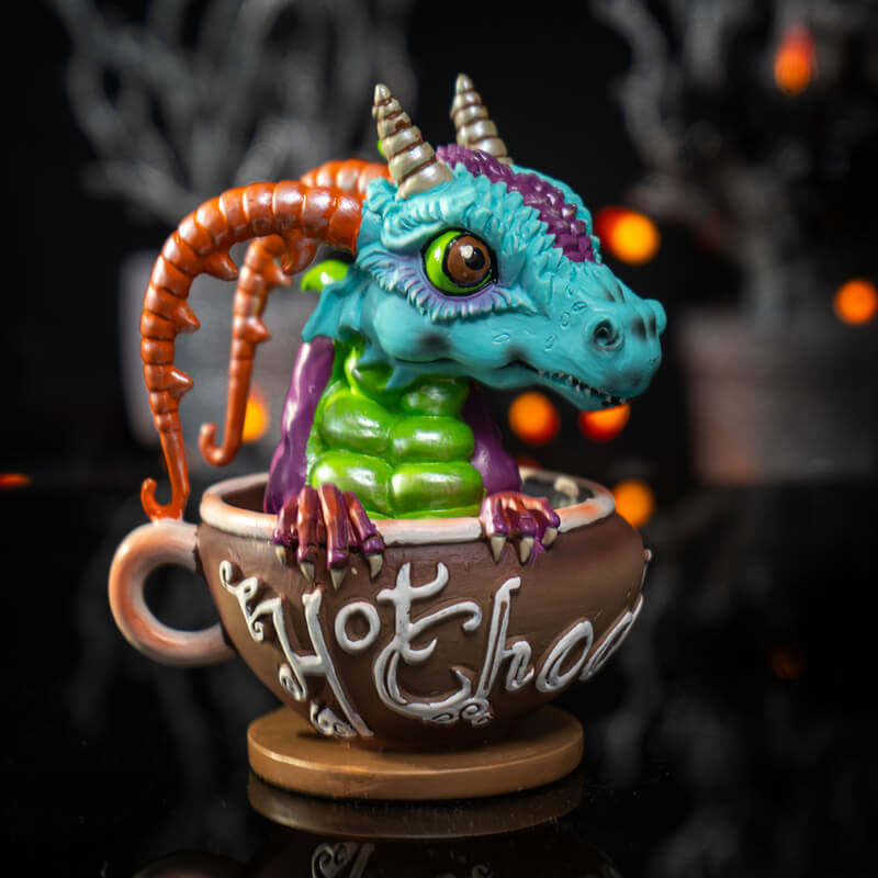Figurine of a blue, green and purple dragon with red horns popping out from a faux mug of hot chocolate with white writing on brown.