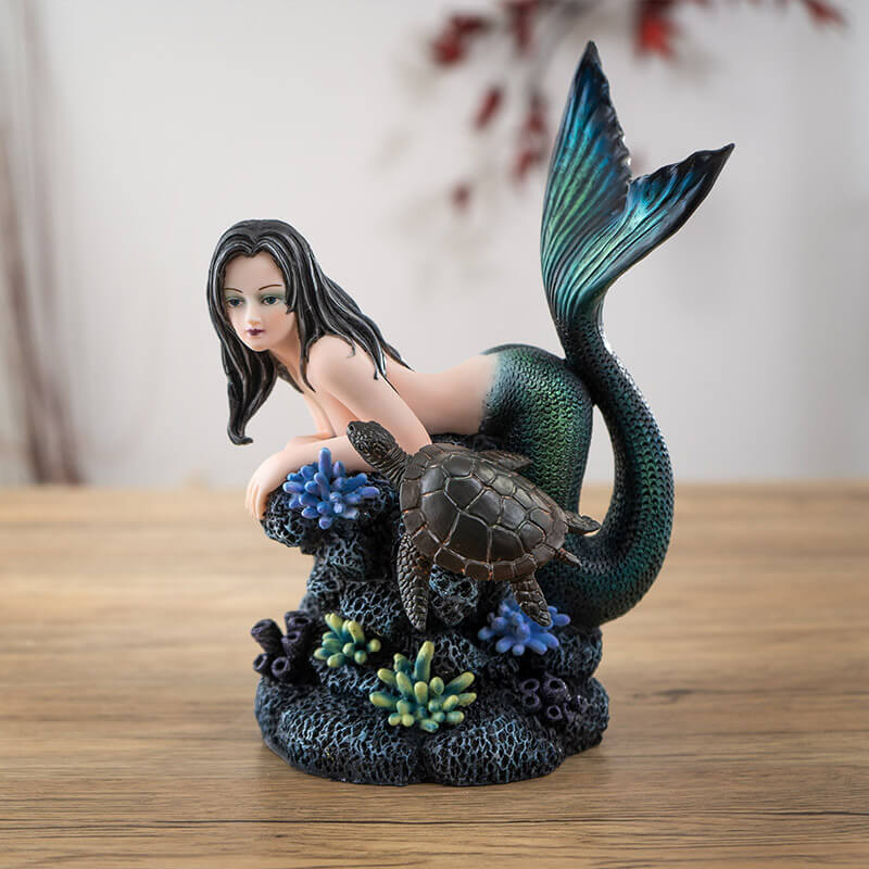 Figurine of black haired mermaid with green and blue tail resting on coral reef with a dark sea turtle