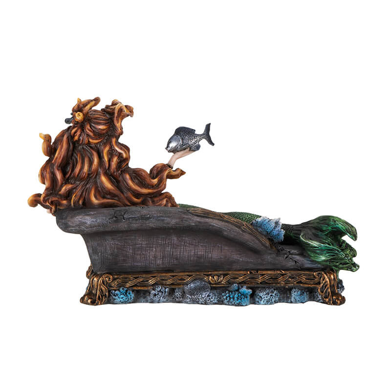 Figurine of a mermaid reclining on a black couch. She has green scales and auburn hair and holds a fish on one hand.