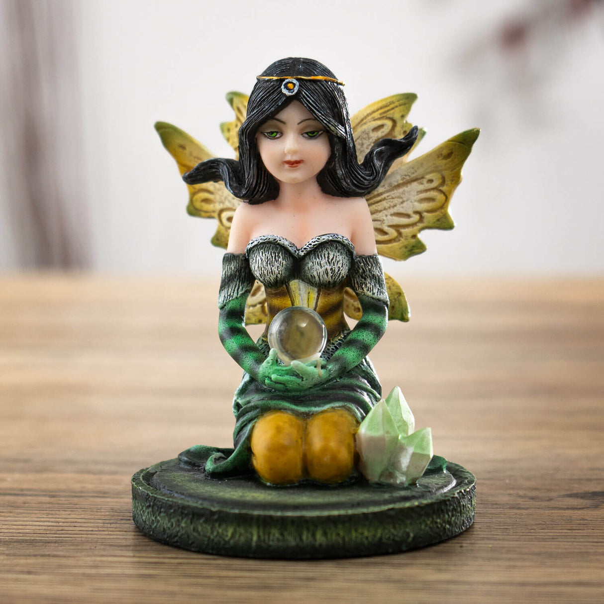 Figurine of kneeling fairy in green, black and yellow with black hair holding a crystal ball on her lap, with crystals next to her
