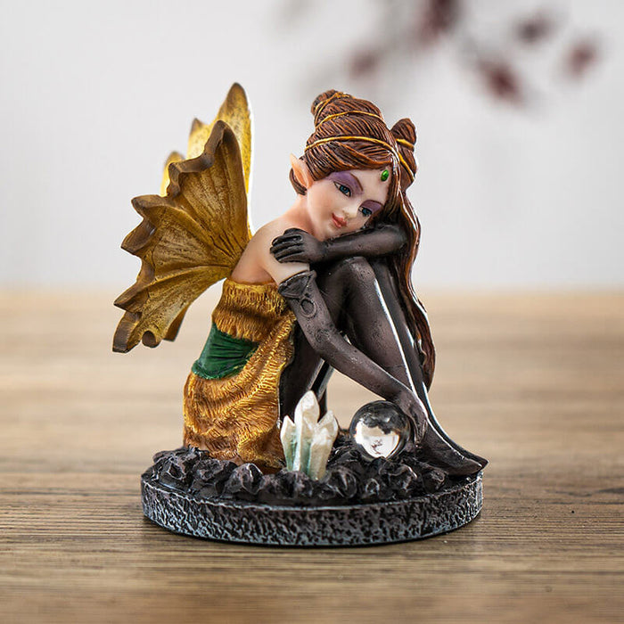 Figurine of a fairy in a gold and green dress with yellow wings, black stockings and gloves. Sitting with a crystal ball and crystal cluster. Elaborate brunette hairdo.