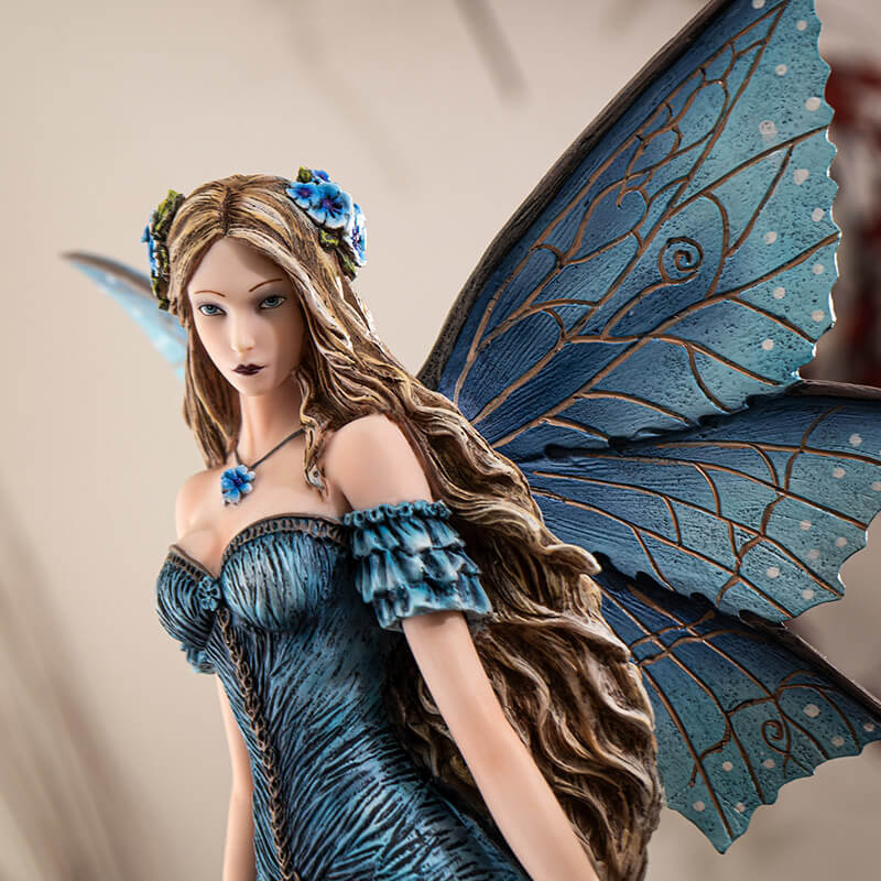 Figurine of a fairy in a blue dress with detailed blue wings and long hair, standing with bright indigo flowers.