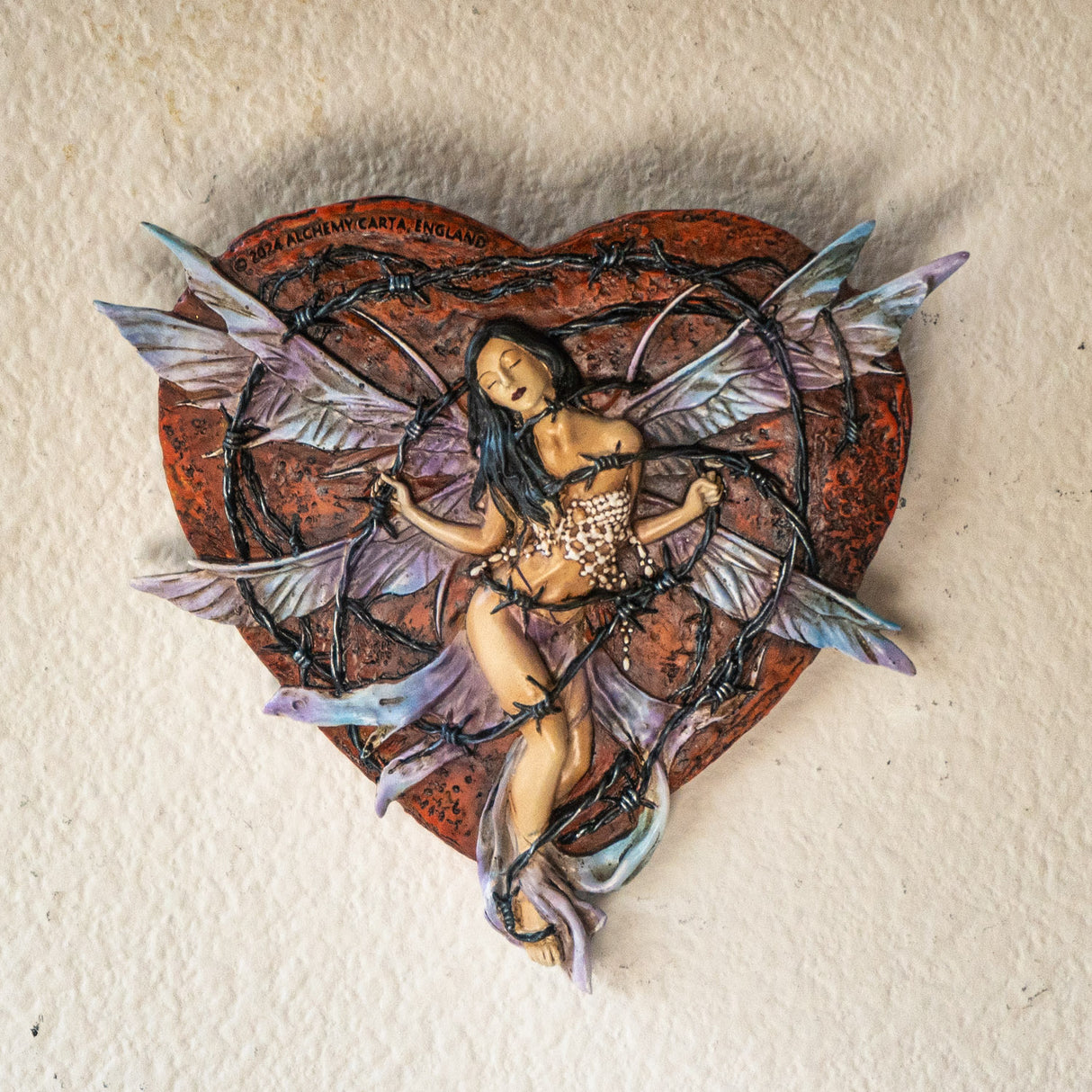 Wall plaque of dark haired fairy with light purple wings trapped in barbed wire against a red heart