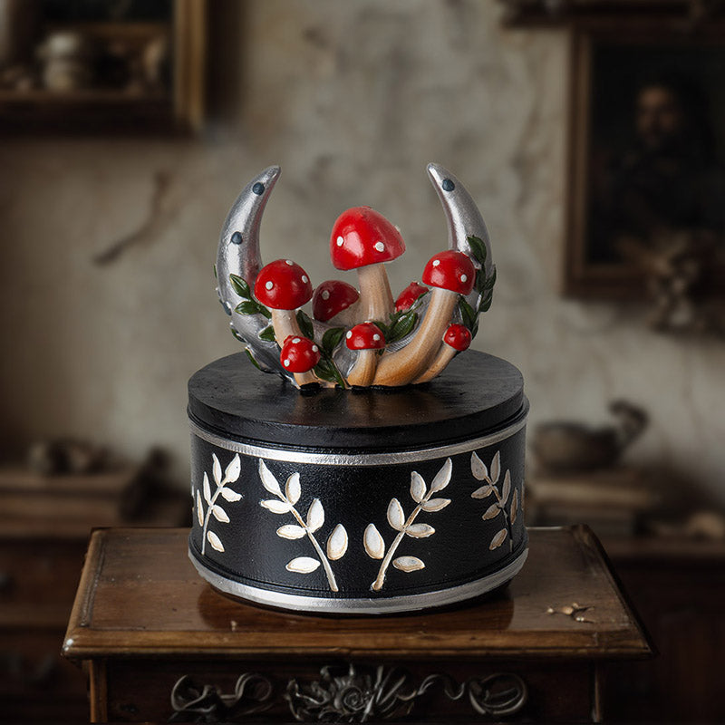 Trinket box with silver moon and red and white mushrooms on the lid, sprigs of leaves around outside of bottom of vessel in white on black.