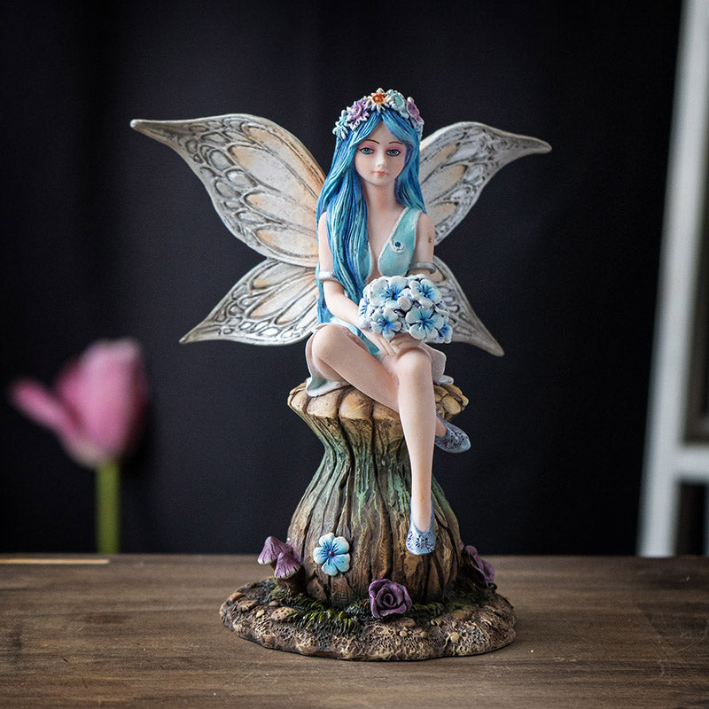 Figurine of fairy with blue hair and tan wings holding blue flower bouquet, sitting on mushroom