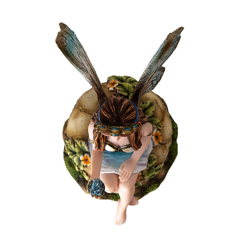 Figurine of a brunette fairy with blue tinged wings and dress sitting on tan mushroom with flowers and leaves, holding baby blue dragon in egg in one palm, shown top down