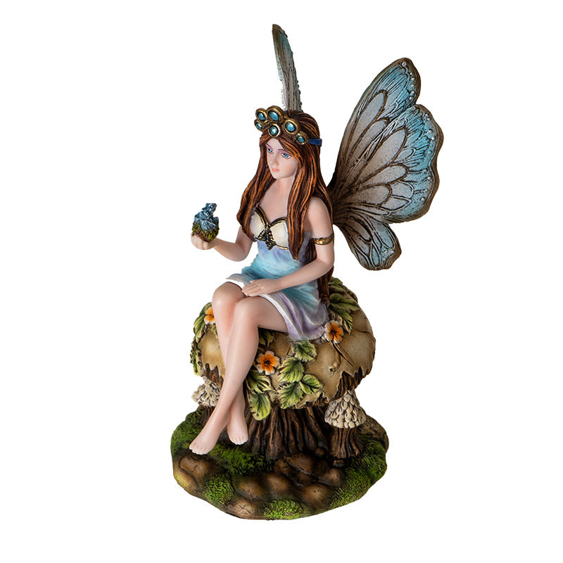 Figurine of a brunette fairy with blue tinged wings and dress sitting on tan mushroom with flowers and leaves, holding baby blue dragon in egg in one palm