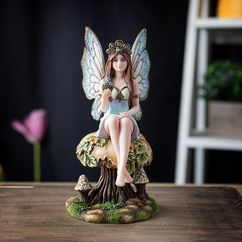Figurine of a brunette fairy with blue tinged wings and dress sitting on tan mushroom with flowers and leaves, holding baby blue dragon in egg in one palm