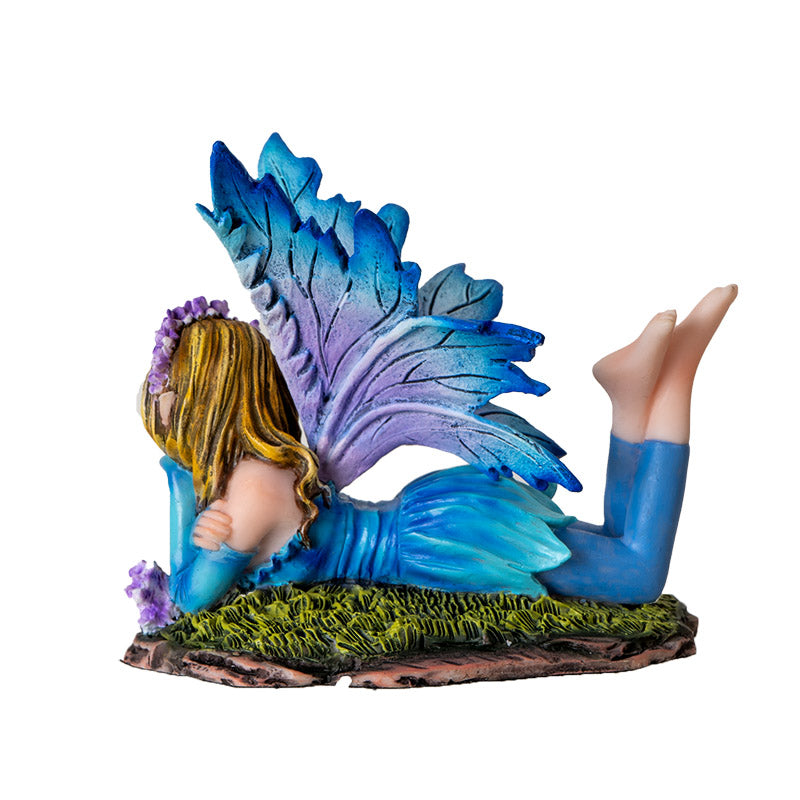 Fairy figurine - pixie laying down, wearing blue dress with leggings and feet kicked up behind her. Blue wings, purple flower and mushroom accents