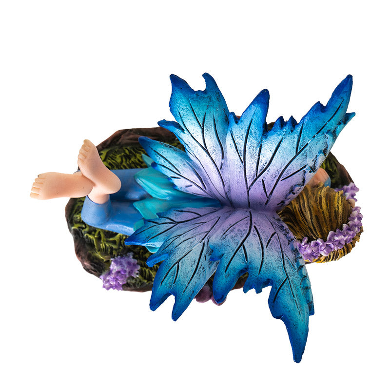 Fairy figurine - pixie laying down, wearing blue dress with leggings and feet kicked up behind her. Blue wings, purple flower and mushroom accents