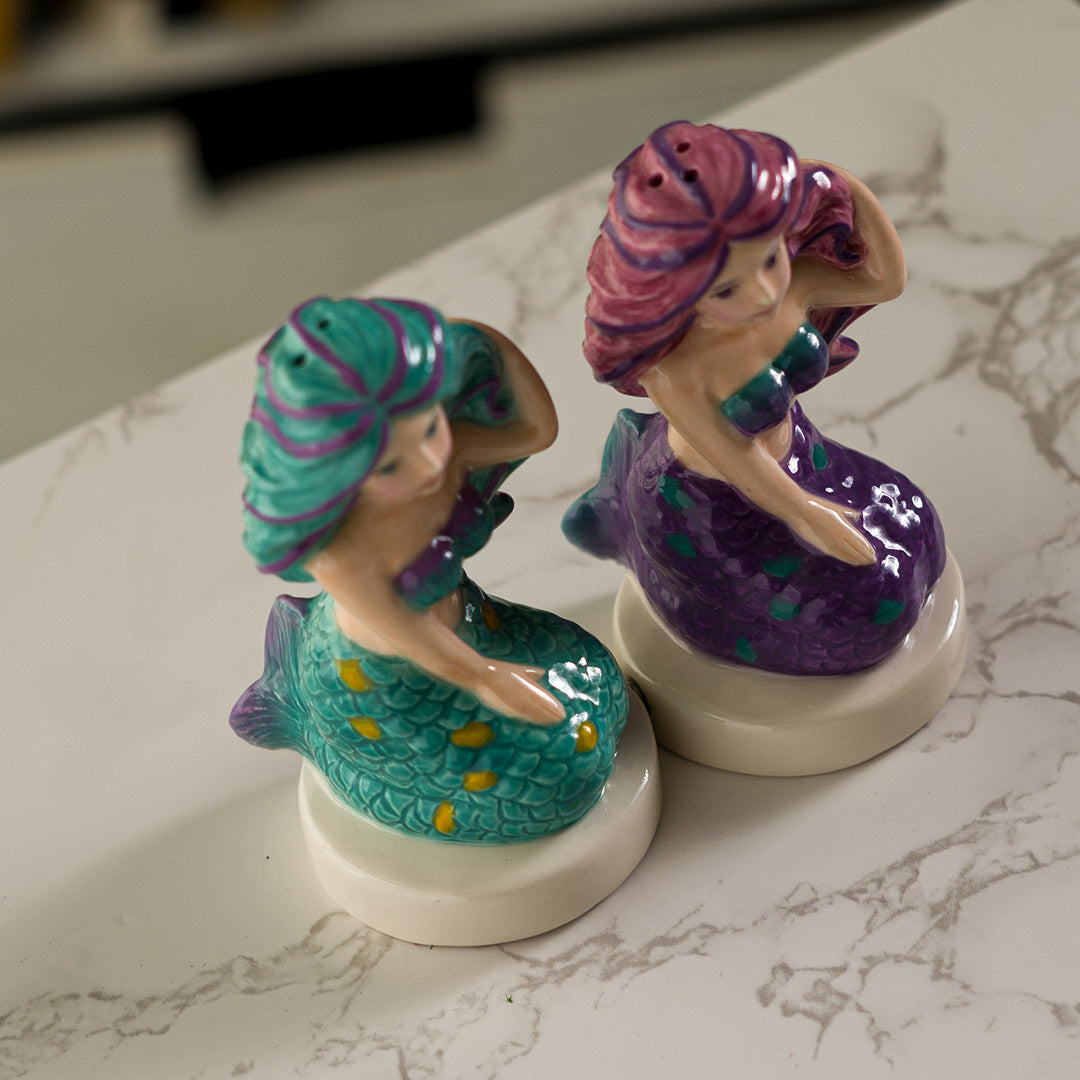 Salt and pepper shakers shaped like mermaids. One in teal with purple and gold accents, one in purple with pink and teal accents