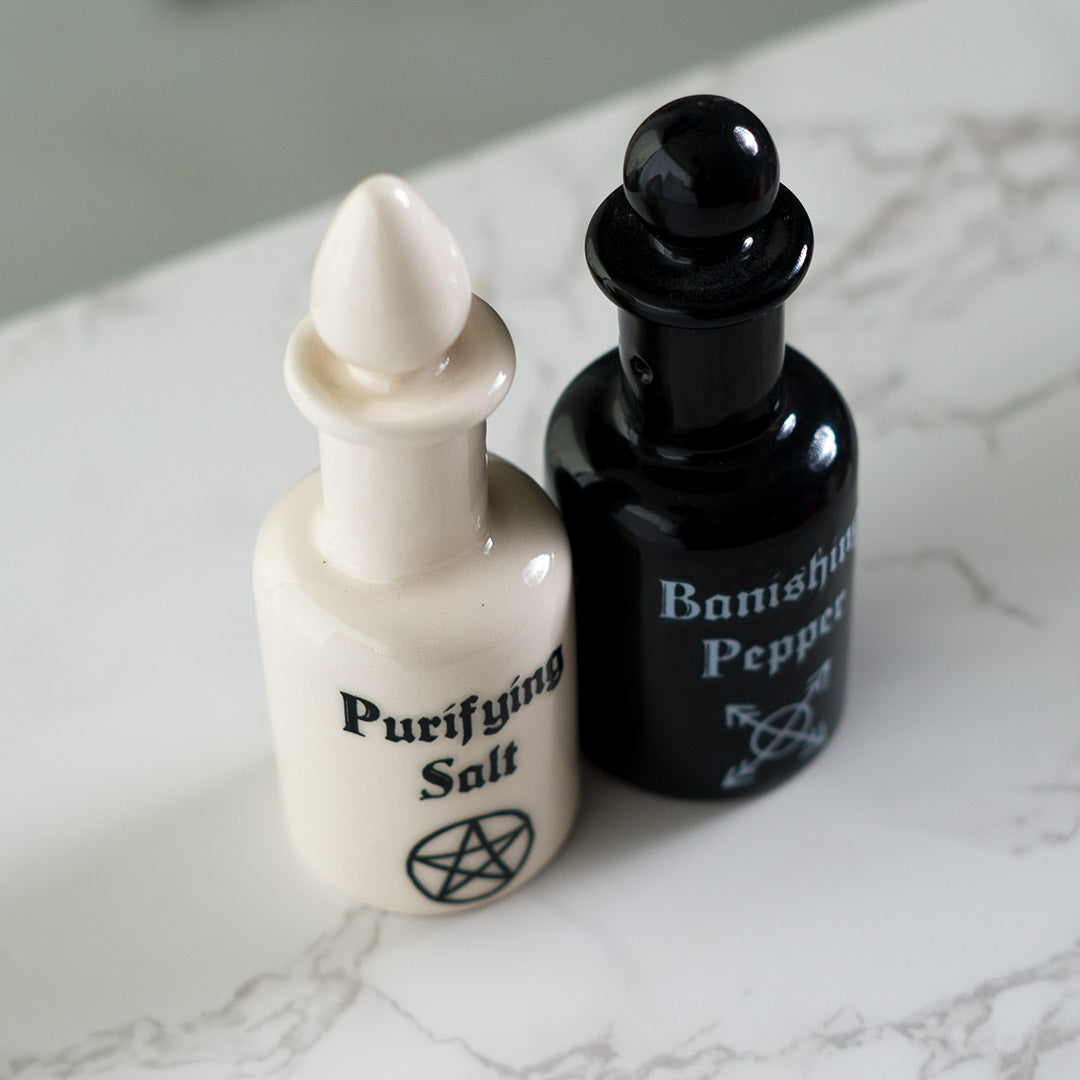 Salt and pepper shakers shaped like potion bottles. White one reads "Purifying Salt" and black one has "Banishing Pepper" with arcane symbols, attached via magnets to one another