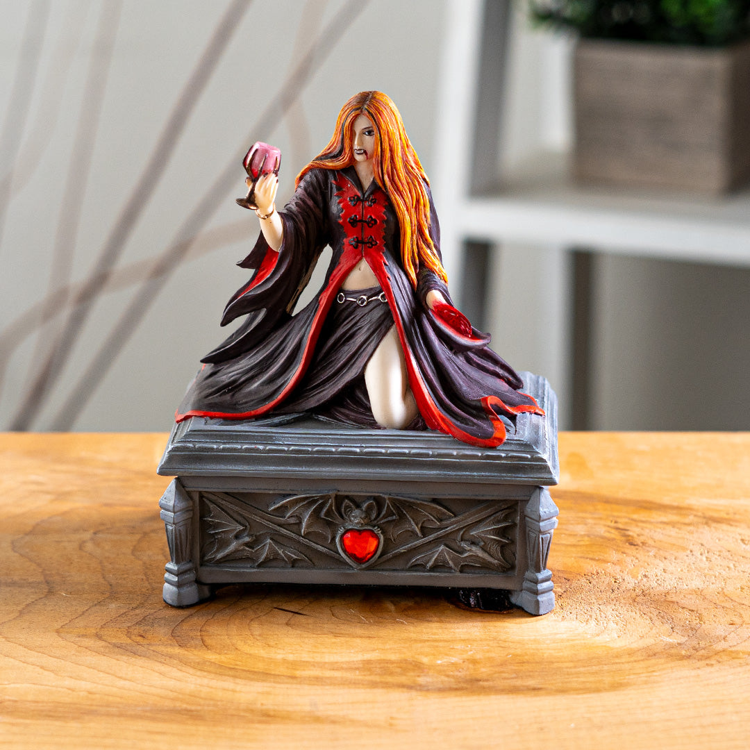 Trinket box, faux stone gray coffin with bat motif and red heart jewel. On lid is a vampire lady with 1 bloody hand and other holding a goblet, crimson and black skirt and top, auburn hair, fangs.