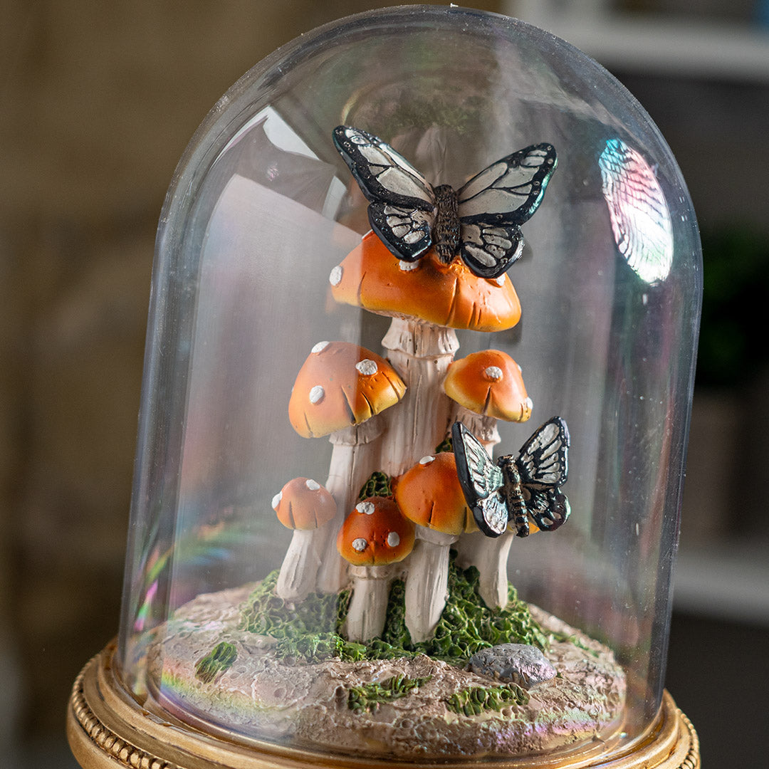 Figurine of butterflies on orange-red mushrooms underneath the glass dome of a cloche, on an ornate golden base
