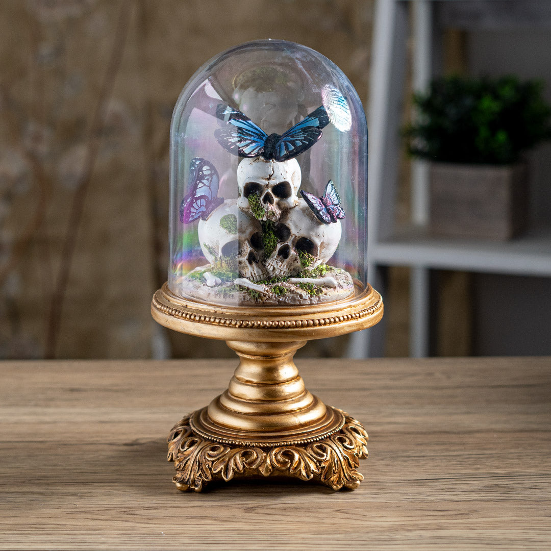 Figurine with three skulls and blue-black butterflies perched on them, moss accents, under a glass cloche dome on an ornate gold base