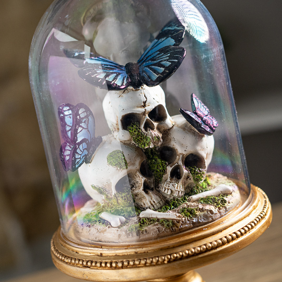 Figurine with three skulls and blue-black butterflies perched on them, moss accents, under a glass cloche dome on an ornate gold base