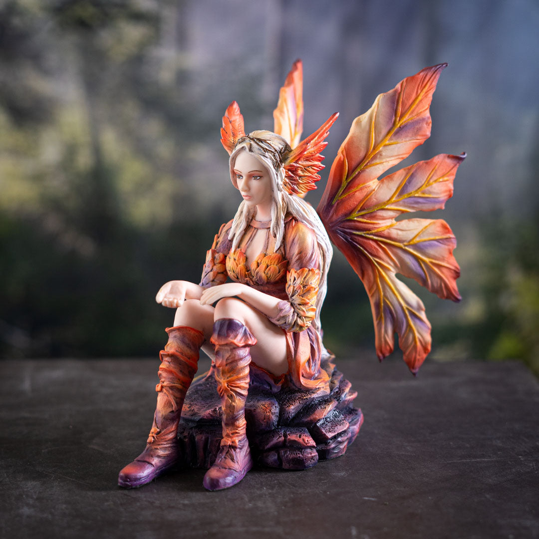Figurine of blond fairy with orang wings and outfit sitting on a rock with her hand out, palm up