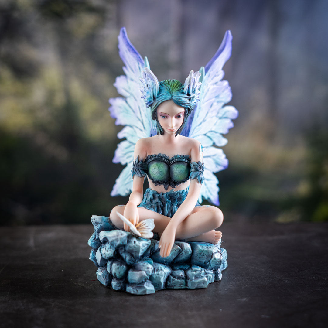Figurine of a sitting fairy, blue-purple wings and green-blue hair, sitting on rocks with a butterfly