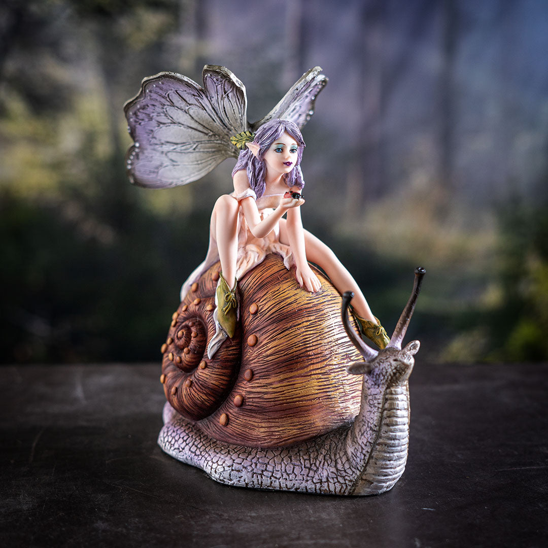 Figurine of a fairy with purple-gray wings and hair, cream colored dress, holding a ladybug and riding a snail