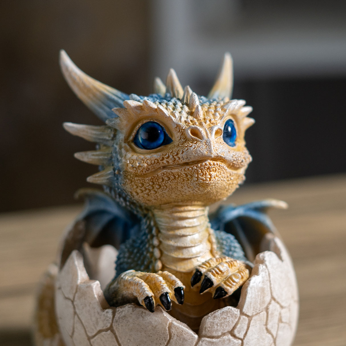A small fantasy dragon figurine with blue scales and bright blue eyes, emerging from a cracked eggshell on a wooden surface with a blurred background.