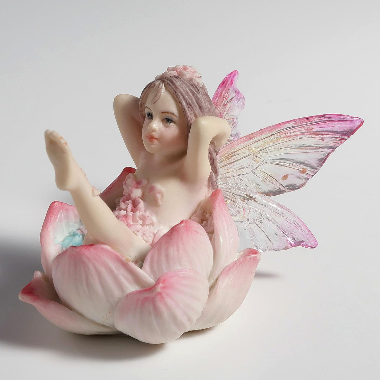 Figurine of a fairy with transparent wings in pink and clear reclining in a flower to have a bath, covered in pink petals