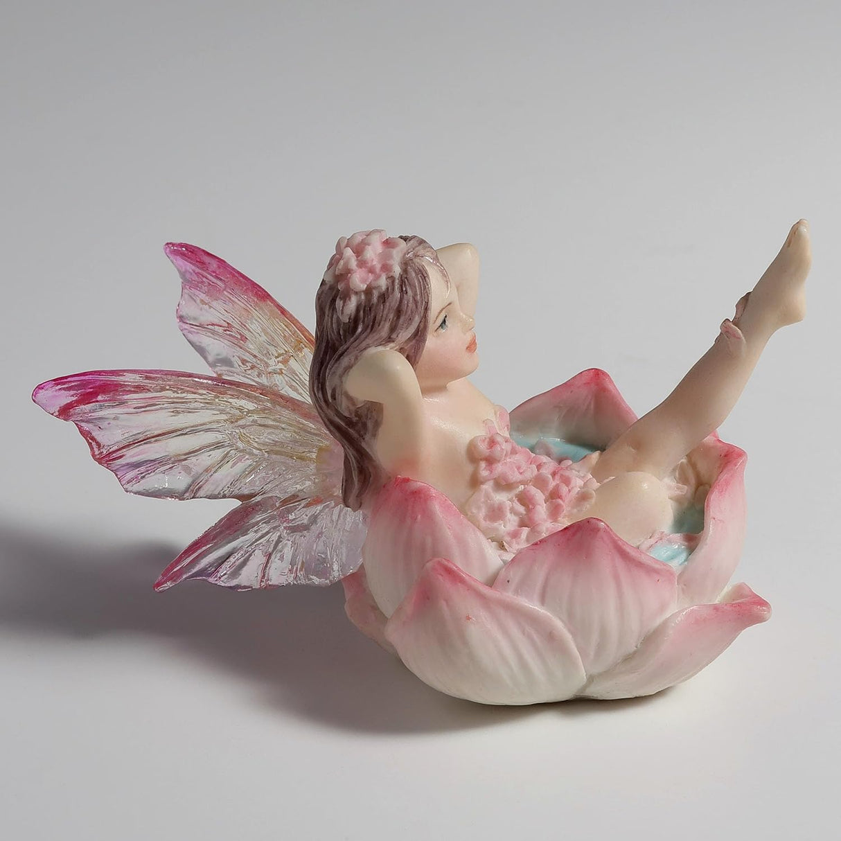 Figurine of a fairy with transparent wings in pink and clear reclining in a flower to have a bath, covered in pink petals