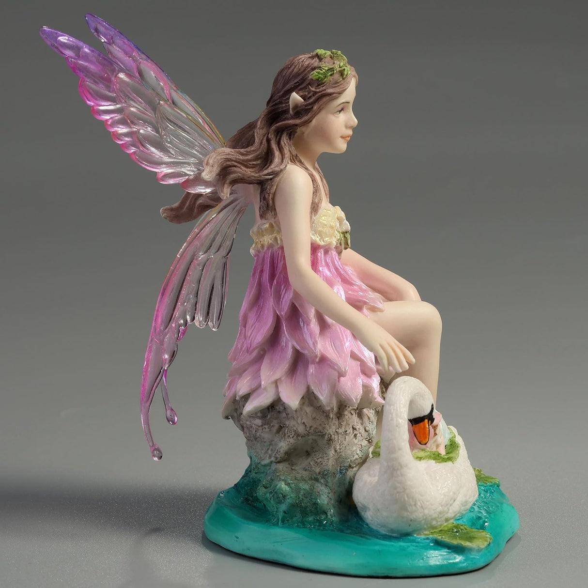 Fairy figurine with pink dress and wings sitting next to a swan with a baby on its back, surrounded by water lilies.