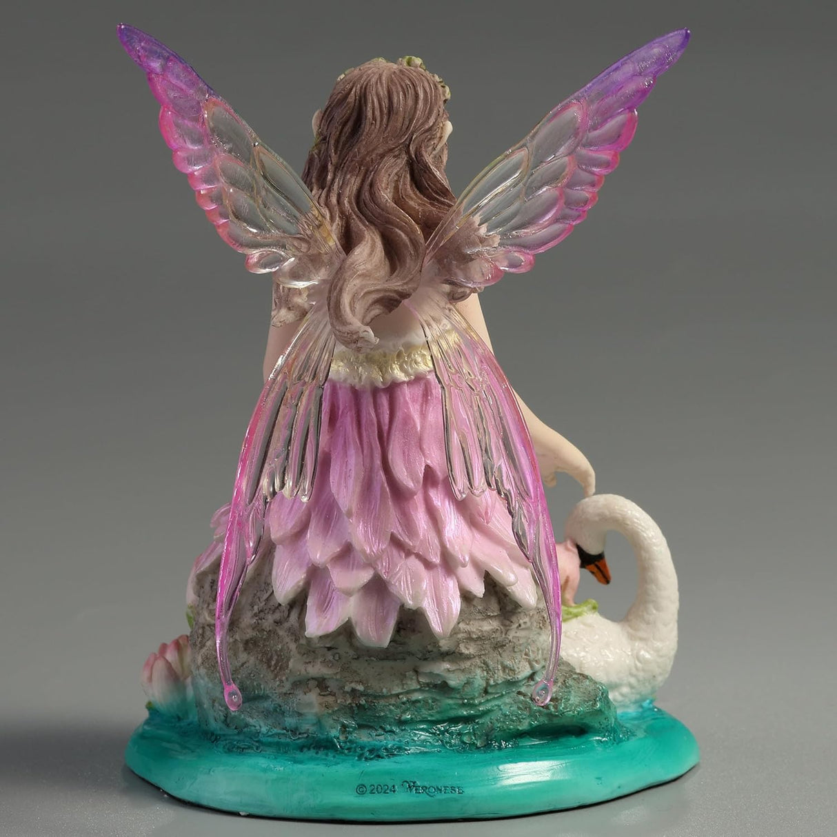 Fairy figurine with pink dress and wings sitting next to a swan with a baby on its back, surrounded by water lilies.