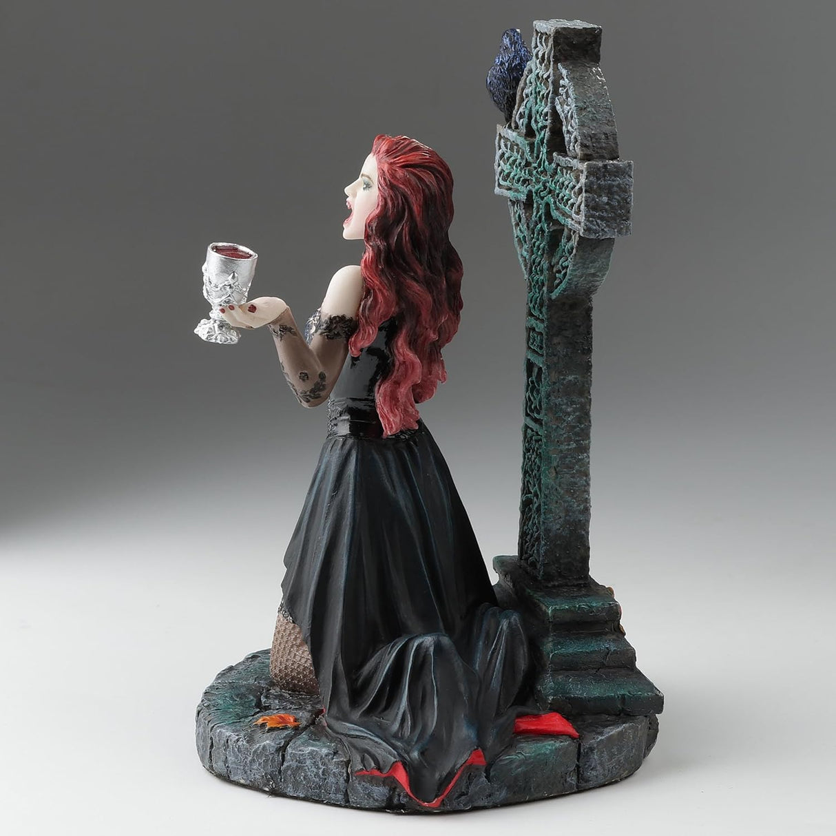 Gothic figurine of a vampire woman with red hair holding a goblet in front of a Celtic cross with a raven perched upon it, shown from the side