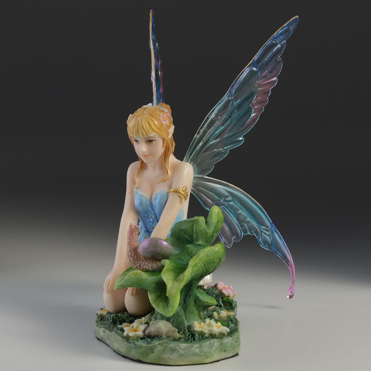 Blond fairy figurine with colorful wings sitting on flowers and grass with a snail on a leaf next to her