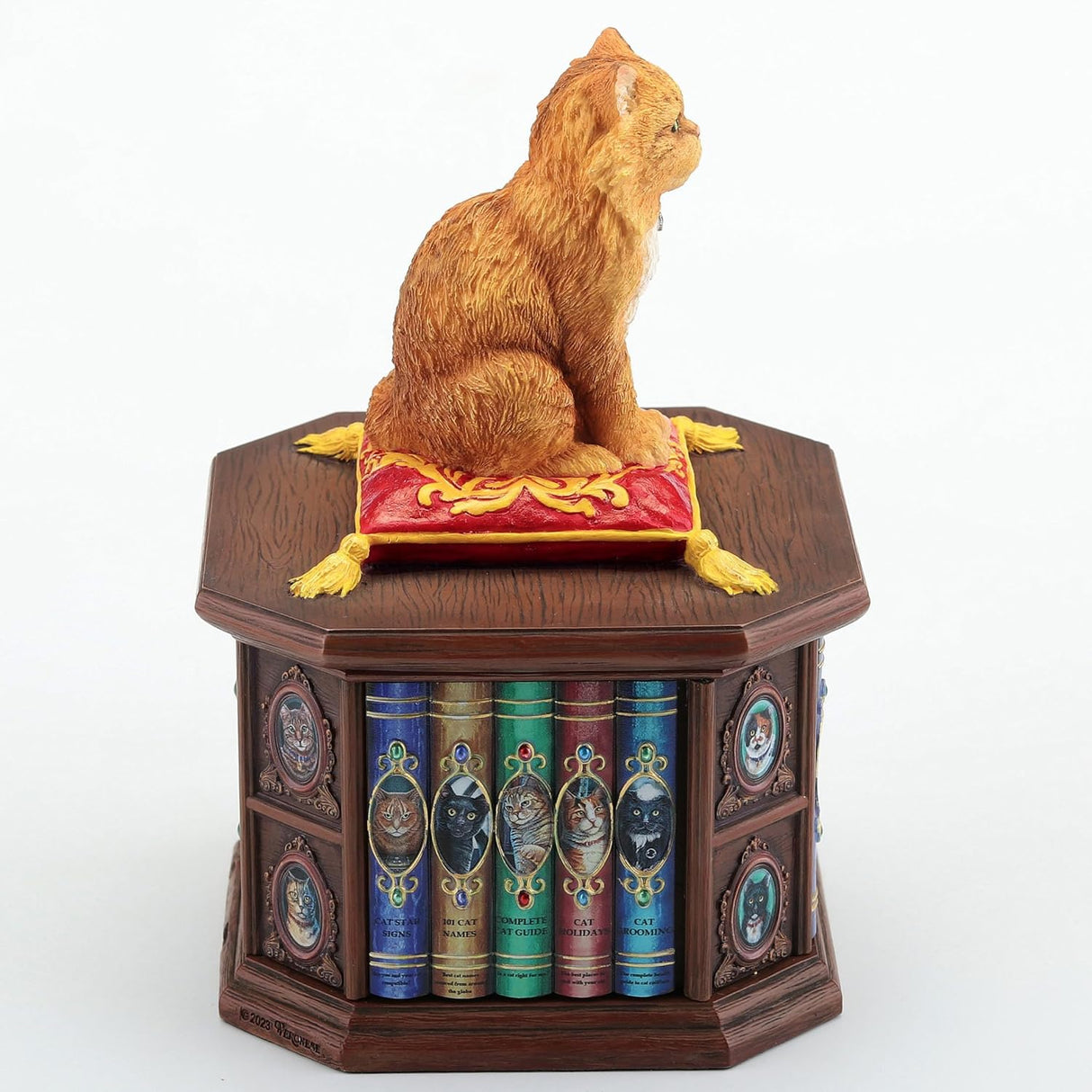 Trinket box of orange cat sitting on a red and yellow pillow on the lid of a faux-wood box shaped like a bookshelf with cat books and pictures. Shown from the back