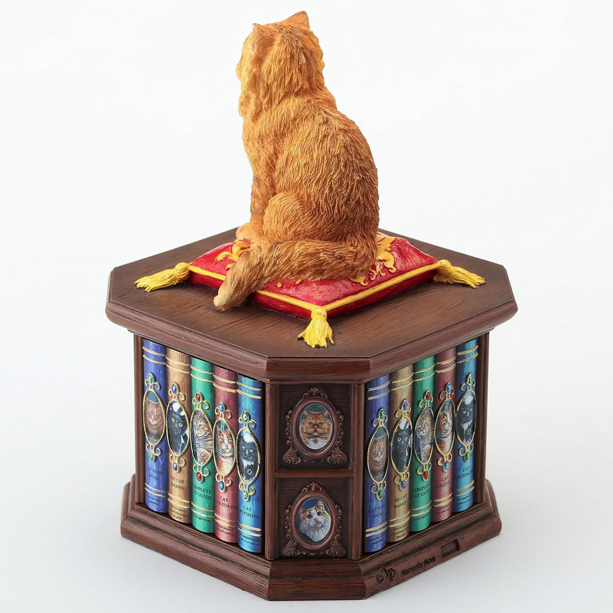 Trinket box of orange cat sitting on a red and yellow pillow on the lid of a faux-wood box shaped like a bookshelf with cat books and pictures. Shown from the back