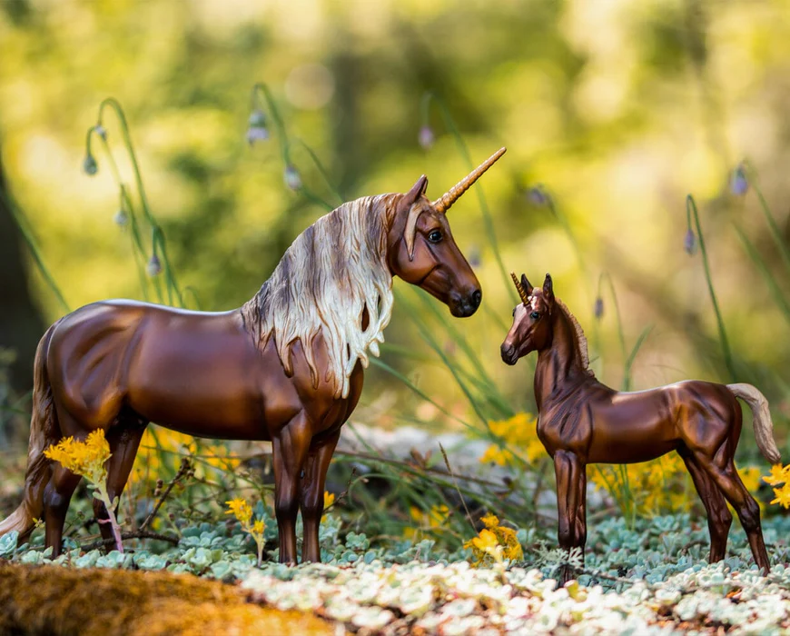 Two brown unicorn figurines with flaxen manes & tails standing on a natural ground with blurred greenery in the background.