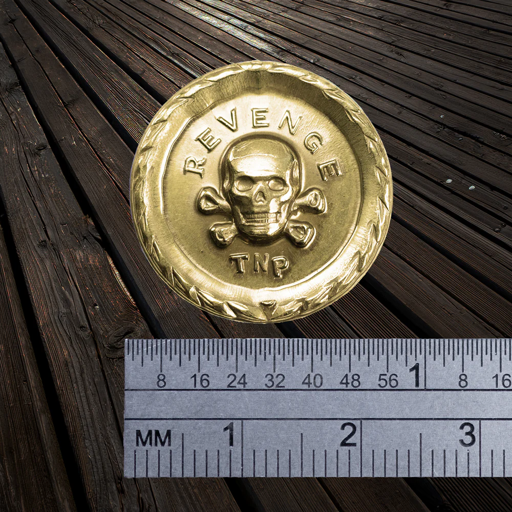 Shiny brass coin with 'REVENGE' and skull design on a wooden surface with a ruler for scale.