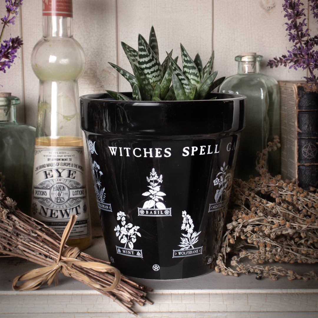 Black ceramic plant pot with the text "Witches Spell Garden" at the top. There are images of herbs including Basil, Mint, Wolfsbane and more, and pentacle designs.
Shown with plants in a witchy kitchen setting