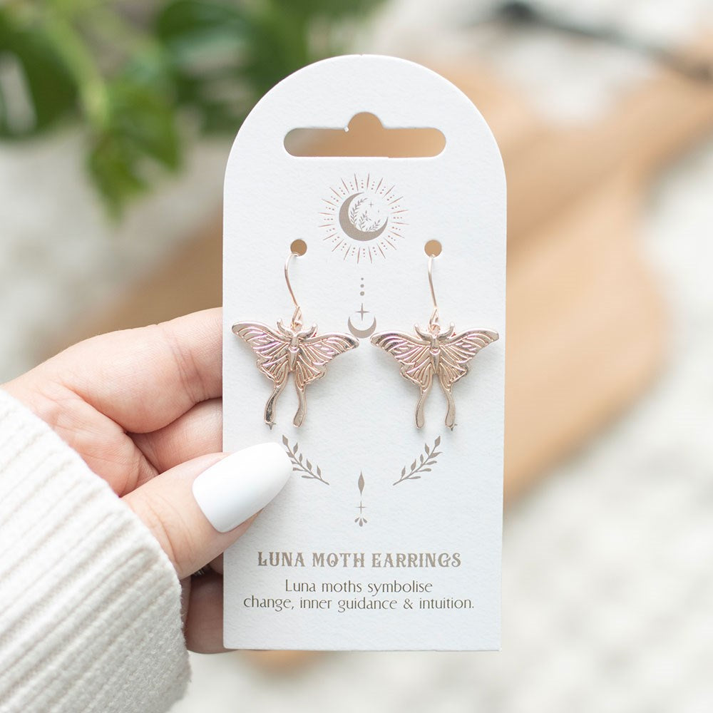 Moth earrings (metal) on backing card with celestial designs and text
"Luna Moth Earrings - Luna moths symbolise change, inner guidance, & intuition." being held by a hand with white nail polish