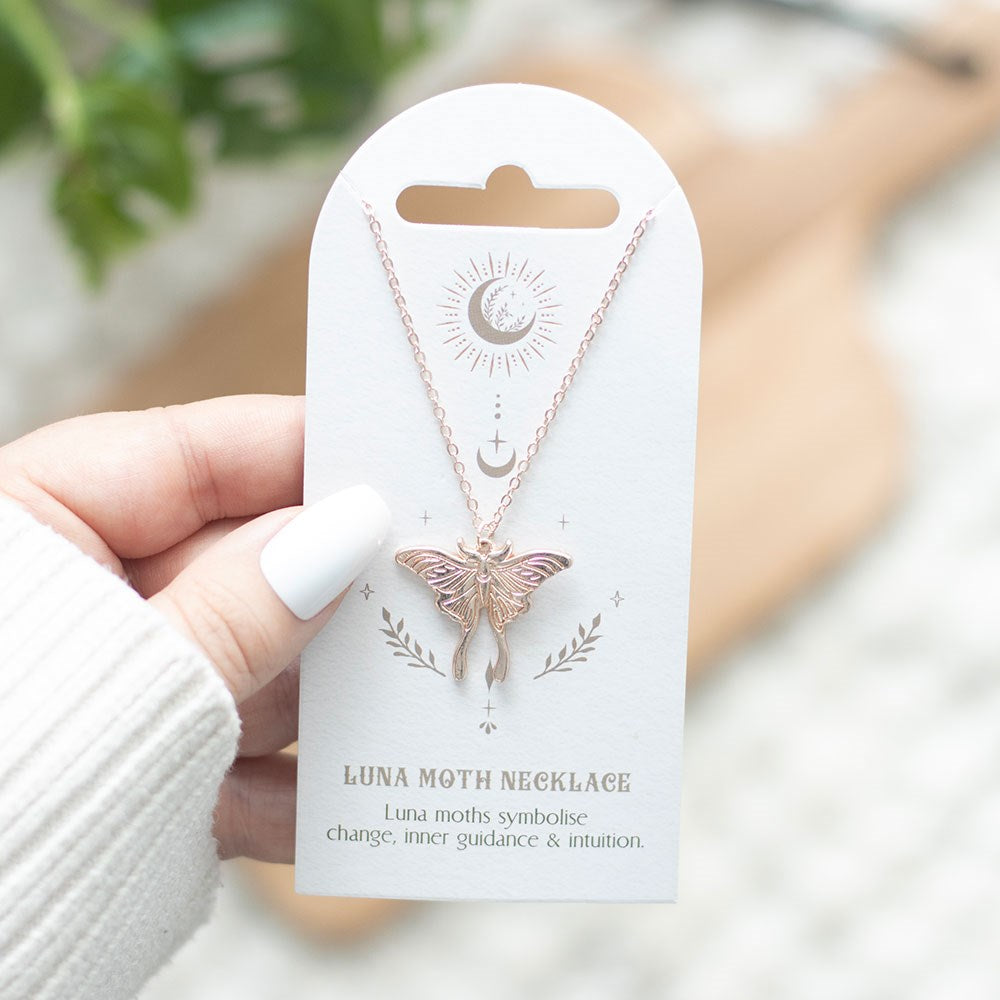 Metal luna moth necklace in a pale bronze-gold color on backing card that reads "Luna Moth Necklace - Luna moths symbolise change, inner guidance & intiution." being held by a hand with white nail polish & sweater