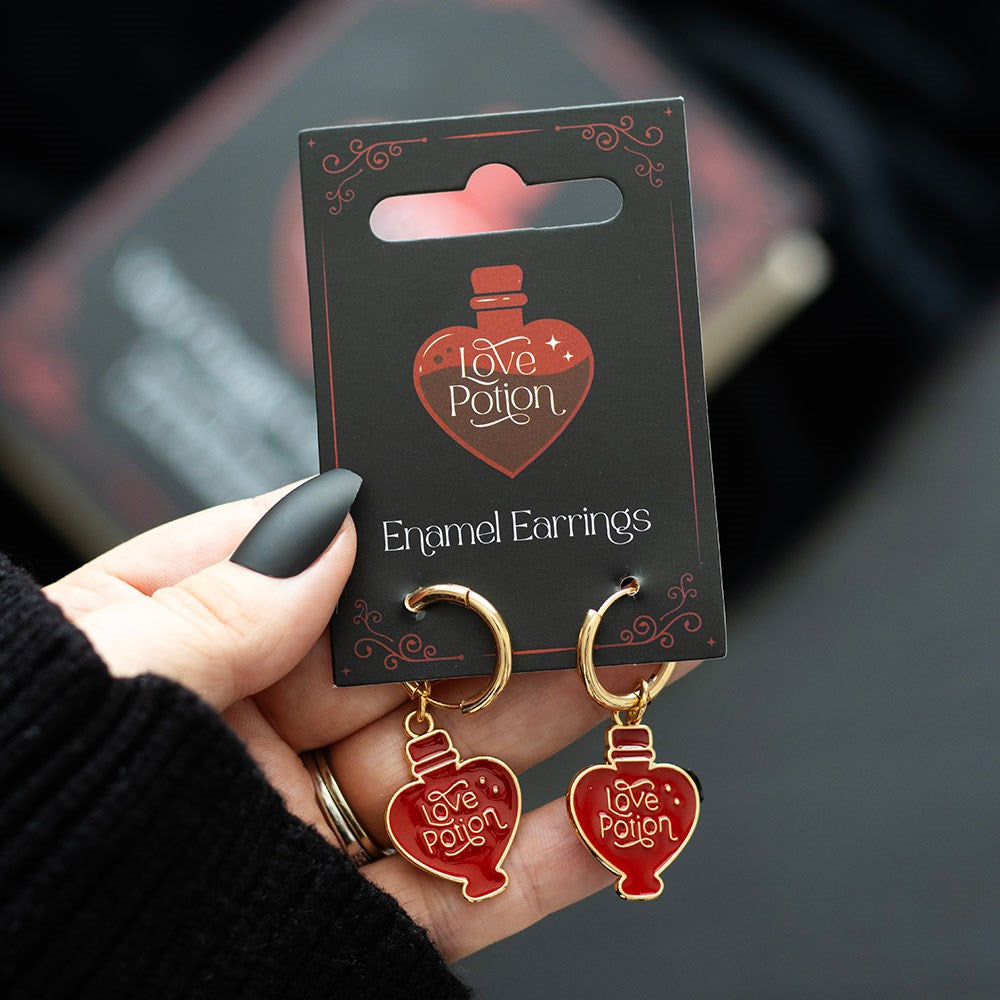 Earrings with red enamel Love Potion heart shaped bottles, gold hoops, shown on their backer card held by hand with black nail polish