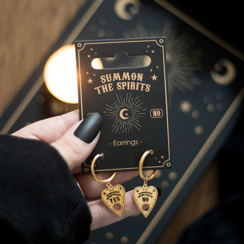 Gold toned ouija talking board planchette earrings on hoops; one says Yes and the other says No with alphabet, stars and moon. Backing card says "Summon The Spirits earrings" and is held by hand with black nailpolish