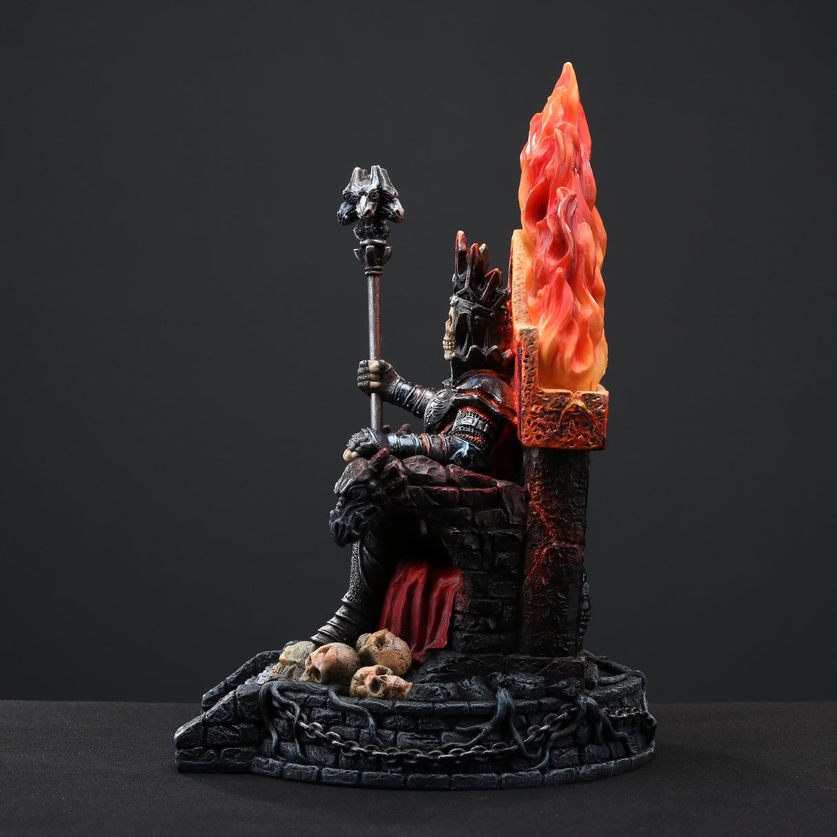 Figurine of skeleton in armor and crown holding staff, sitting on fiery throne with gargoyles, piles of human skulls, shown from the side