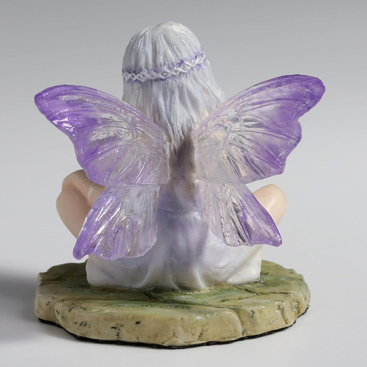 Figurine of fairy with purple-clear wings and pale silver hair sitting cross legged on a mossy stone, shown from the back
