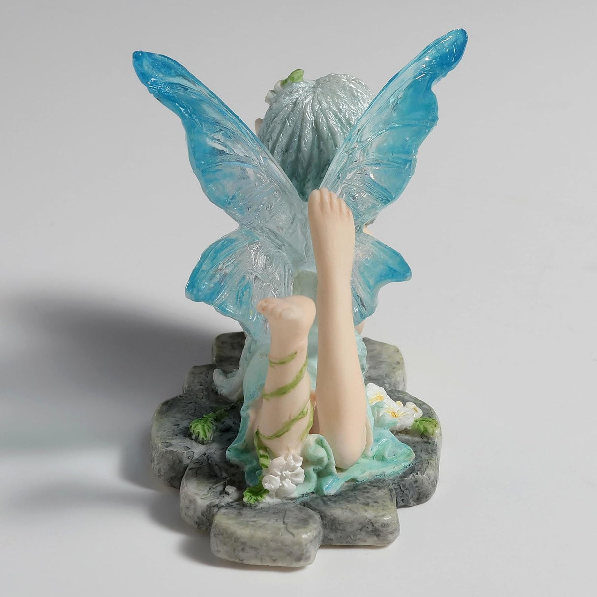 Figurine of fairy in pale blue dress with clear and blue wings and braided hair lying amidst flowers on a stone, feet kicked up