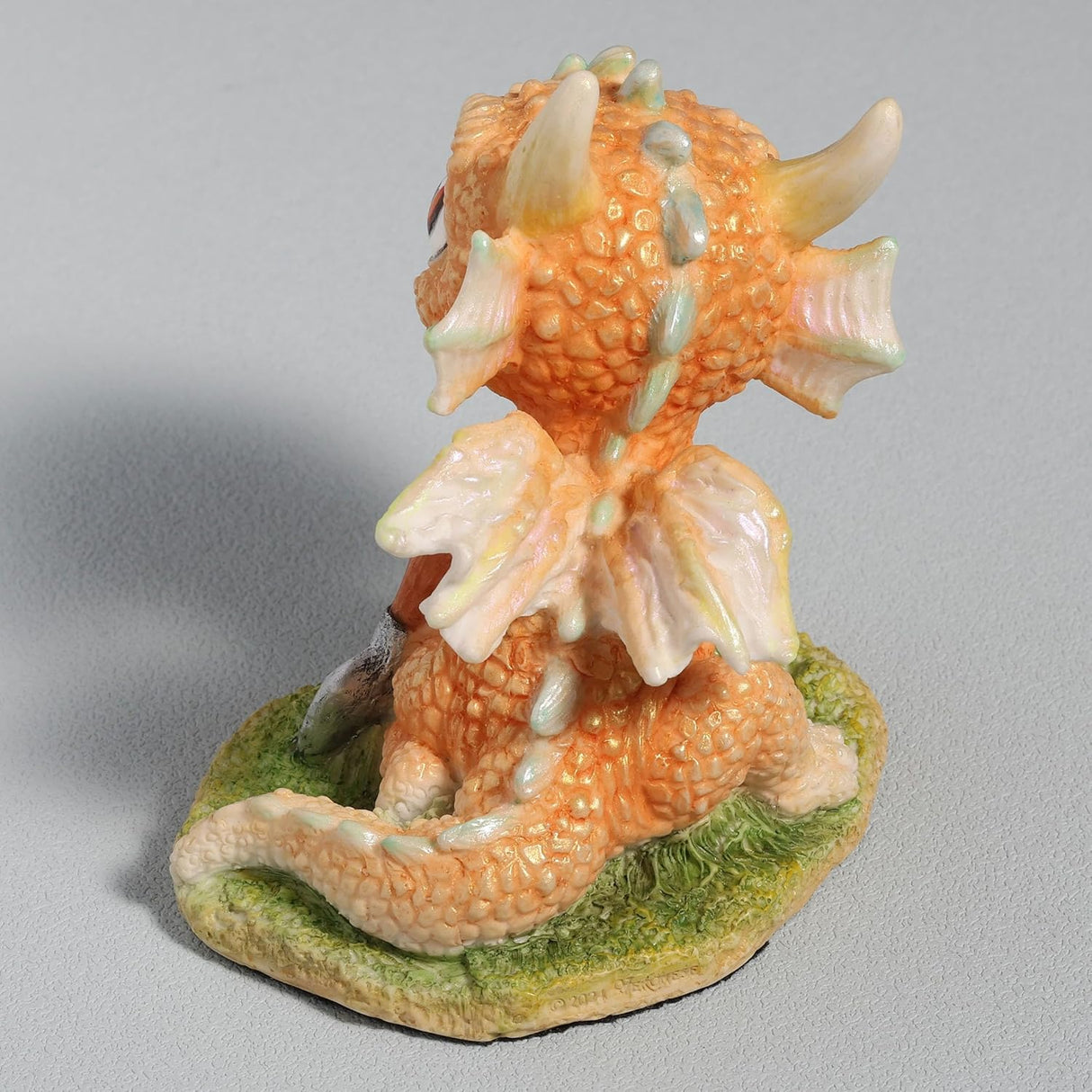 Figurine of a pale gold baby dragon gardening with flowers and a trowel, shown from the back