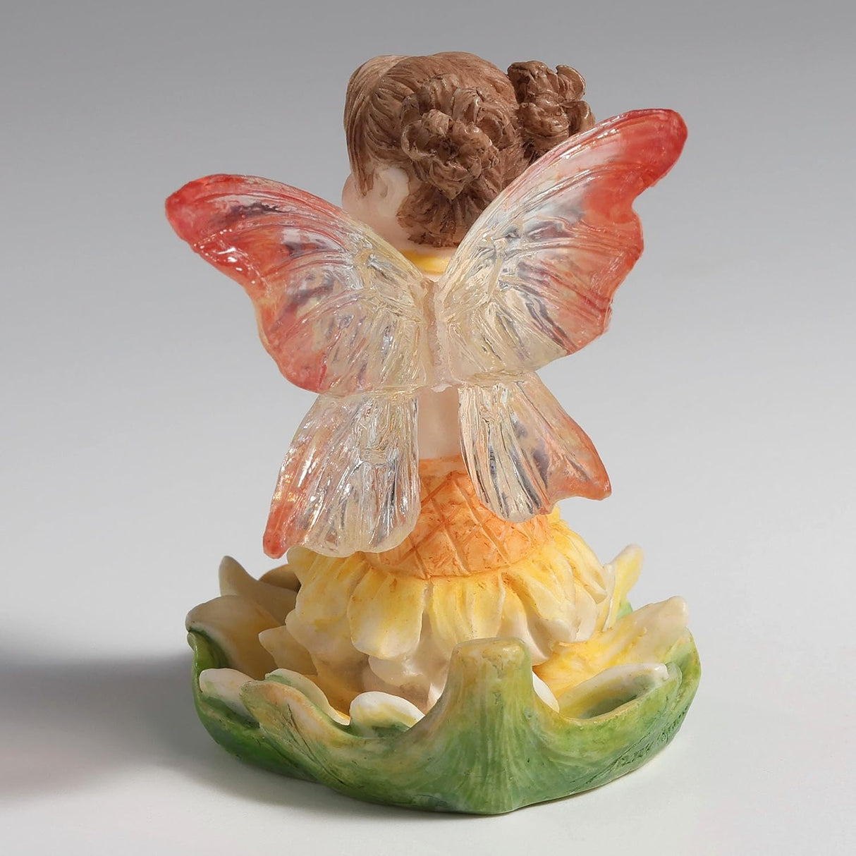 Figurine of fairy in yellow-orange dress with pink and clear wings, reddish-brown hair, blue eyes sitting in a leaf, shown from the back