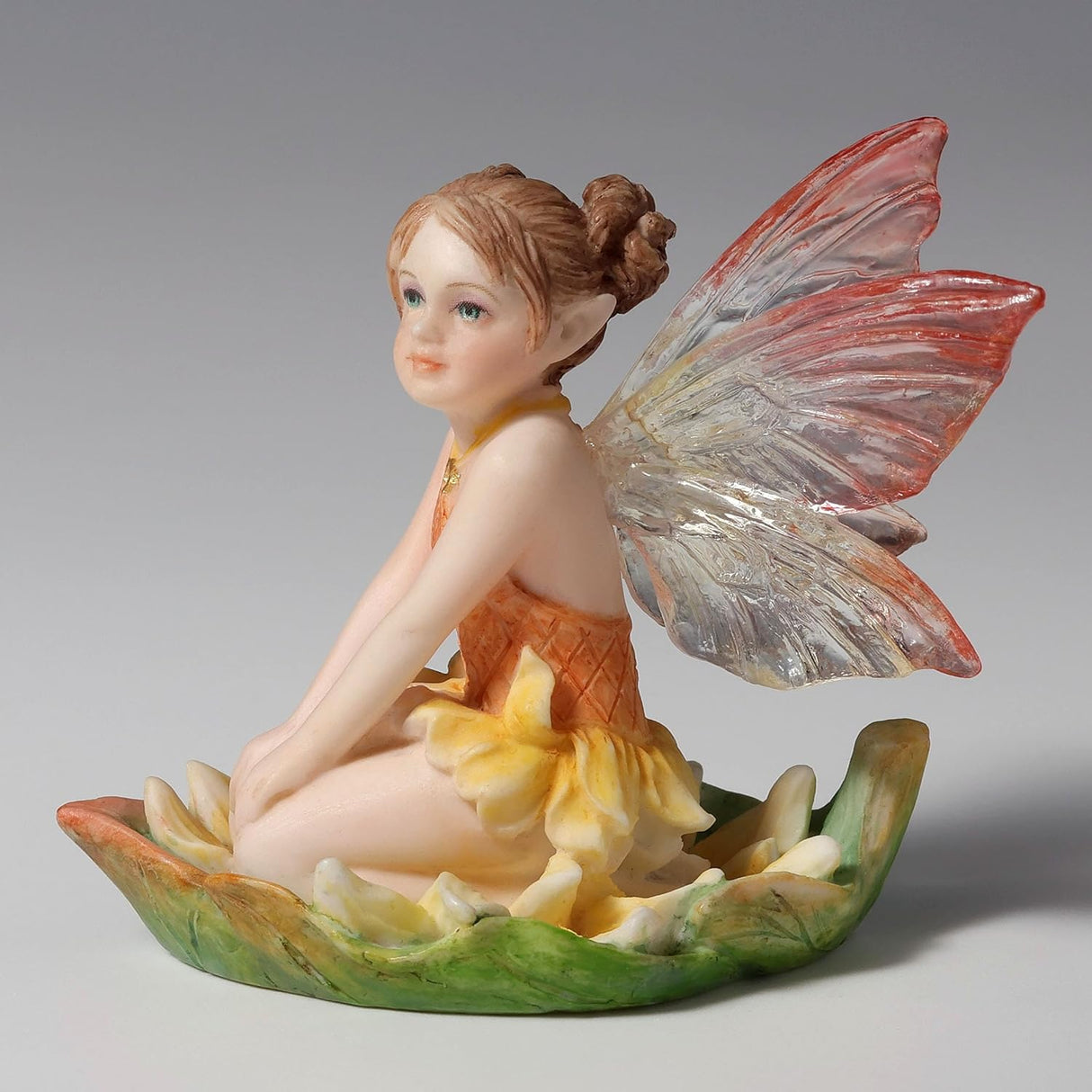 Figurine of fairy in yellow-orange dress with pink and clear wings, reddish-brown hair, blue eyes sitting in a leaf