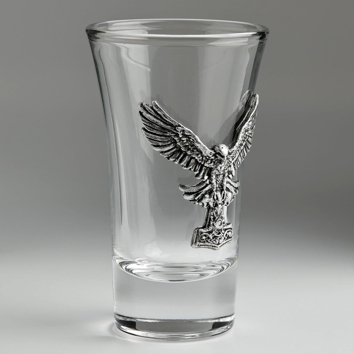 A clear glass shot glass featuring a detailed, raised pewter hawk emblem holding a hammer on the front